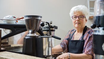 Ältere Frau mit Schürze bedient eine Kaffeemaschine | © tyler_olson_60009761